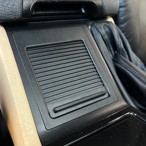 Puede incluir: Primer plano del interior de un coche negro con un reposabrazos de cuero beige y un compartimento de almacenamiento de plástico negro con una ventilación.