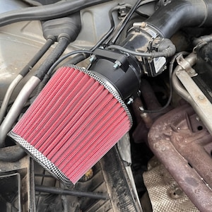 Puede incluir: Un filtro de aire de alto rendimiento rojo y negro con un tubo de admisión negro. El filtro está instalado en el compartimento del motor de un automóvil.