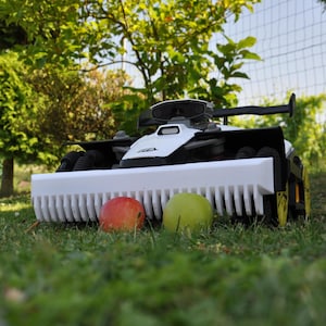 Può includere: Un tosaerba robotico bianco e nero con una struttura a forma di pinna e ruote gialle è su un prato erboso. Una mela rossa e una mela verde sono davanti al tosaerba. Lo sfondo comprende alberi e una recinzione.