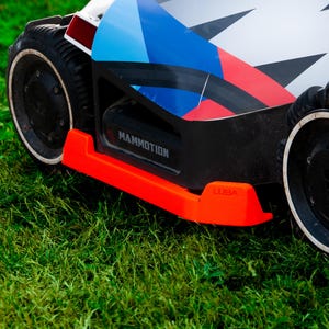 May include: A robotic lawn mower featuring a white, blue, and red geometric graphic on its top surface. The device has black wheels with white rims, a bright orange front bumper, and the text MAMMOTION and LUBA printed on its black chassis.