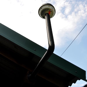 May include: A black, curved pipe with a white and black circular fixture attached to a green roof. The sky is visible in the background, with a few clouds.