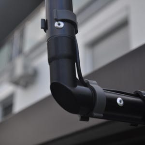 Op de afbeelding: Close-up van een zwarte, gebogen metalen buis met grijze plastic clips en een witte schroef. De buis is in een hoek van 90 graden verbonden met een andere zwarte buis. De achtergrond is wazig en toont een gebouw.