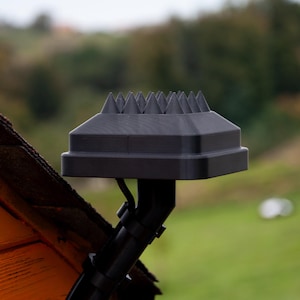 Może przedstawiać: Czarny ochraniacz rynny z kolczastym wierzchem, zaprojektowany w celu zapobiegania gromadzeniu się zanieczyszczeń. Ochraniacz jest zamontowany na czarnej rurze spustowej, z rozmytym zielono-brązowym tłem.