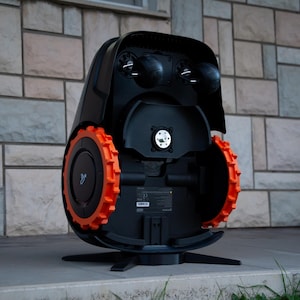 Pode incluir: Cortador de relva robótico preto e laranja com um design moderno. O cortador de relva tem duas rodas grandes laranja e um corpo preto. A parte superior do cortador de relva tem três elementos circulares pretos. O cortador de relva está numa superfície de betão.