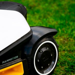以下が含まれることがあります： 緑の芝生の上にある、白と黒のロボット芝刈り機のクローズアップ。芝刈り機は白い上部、黒いボディ、白いリムのホイールを備えています。「MAMMOTION」の文字が見えます。