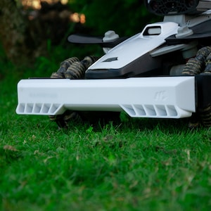 以下が含まれることがあります： 緑の芝生の上にある白と黒のロボット芝刈り機。芝刈り機は、白いフロントバンパーと黒い上部を備えたロープロファイルです。ホイールは暗く、質感があります。芝刈り機は、自動芝生ケア用に設計されています。