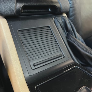 May include: A close-up of a car's interior console. The console is black with a textured, rectangular vent and a small, rectangular storage compartment. The surrounding trim is beige, and the seats are black leather.