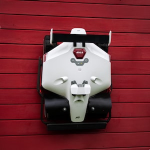 May include: A white and black robotic lawn mower with a red "STOP" button and a black spoiler. The mower is mounted on a red wooden wall. The mower has a futuristic design.