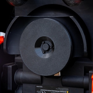 May include: Close-up of a black, circular grinding wheel with a textured surface. The wheel is mounted within a black housing, with a central hub and several visible screws. The overall design suggests a power tool or machine component.