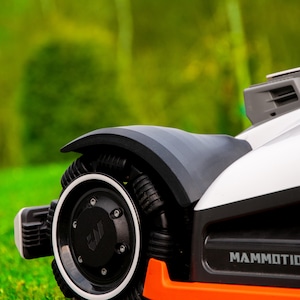 May include: Close-up of a robotic lawnmower with a black wheel and fender, white body, and orange accents. The wheel has a white rim and a black centre with the letters "MP". The lawnmower is on a green lawn.