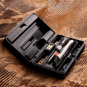 Könnte beinhalten: Geöffnete schwarze Werkzeugkiste mit verschiedenen Werkzeugen, darunter Schraubendreher, eine Bürste und Kleinteile. Auf einer strukturierten braunen Oberfläche.