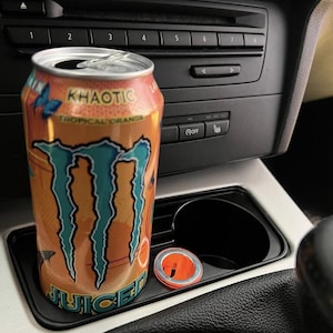 Könnte beinhalten: Eine orangefarbene und türkisfarbene Khaotic Tropical Orange Energy-Drink-Dose befindet sich in einem Getränkehalter im Auto. Auf der Dose stehen die Wörter "Khaotic" und "Juiced" mit einer Schmetterlingsgrafik. Daneben liegt ein kleines orangefarbenes und silbernes Objekt.