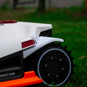 May include: A close-up of a white and black robotic lawn mower with an orange accent. The mower has a black wheel with a white rim and a red light. The word "MMOTION" is printed on the side.