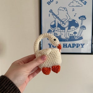 Pu&ograve; includere: Un cigno bianco all'uncinetto con piedi arancioni. Il cigno &egrave; tenuto nella mano di una persona. Lo sfondo &egrave; una parete bianca con un poster incorniciato che dice "Fai ci&ograve; che ti rende felice" con una rana di cartone animato che suona il banjo.