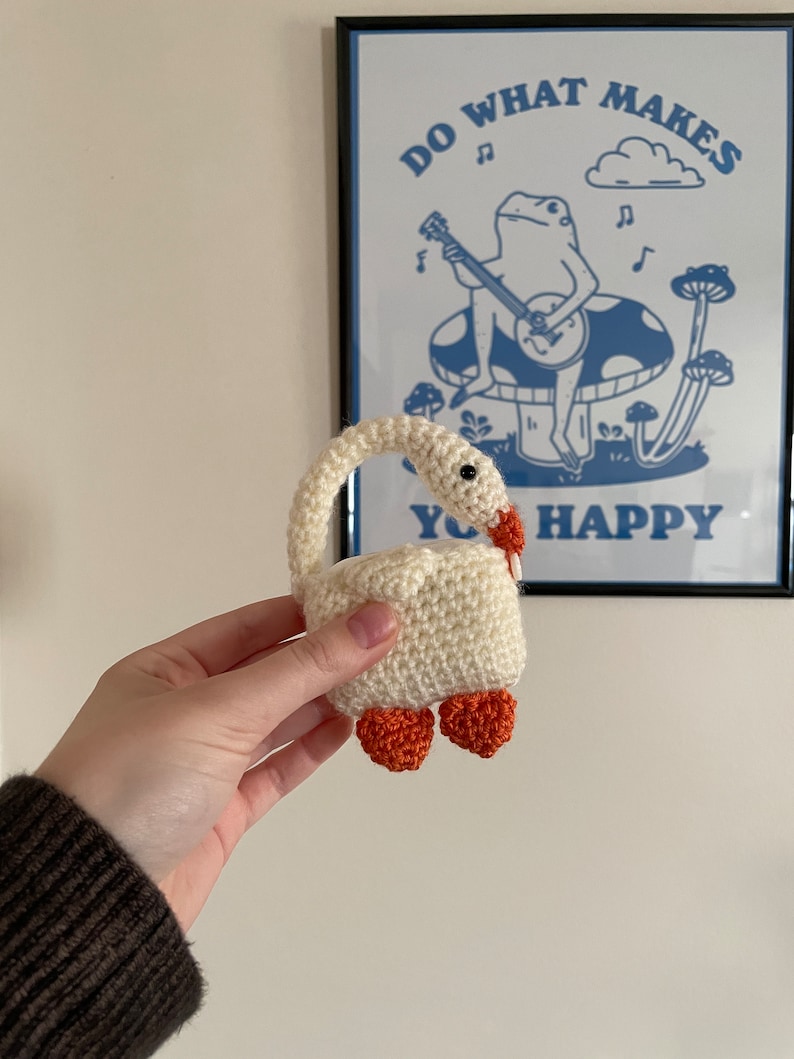 Pu&ograve; includere: Un cigno bianco all'uncinetto con piedi arancioni. Il cigno &egrave; tenuto nella mano di una persona. Lo sfondo &egrave; una parete bianca con un poster incorniciato che dice "Fai ci&ograve; che ti rende felice" con una rana di cartone animato che suona il banjo.