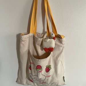 Pu&ograve; includere: Una borsa tote in tela beige con cinturini gialli. La borsa ha una tasca frontale con un'illustrazione di un pollo che indossa un cappello di fragola e il testo "berry time". Un piccolo anatroccolo bianco all'uncinetto con dettagli rossi &egrave; appeso alla borsa.