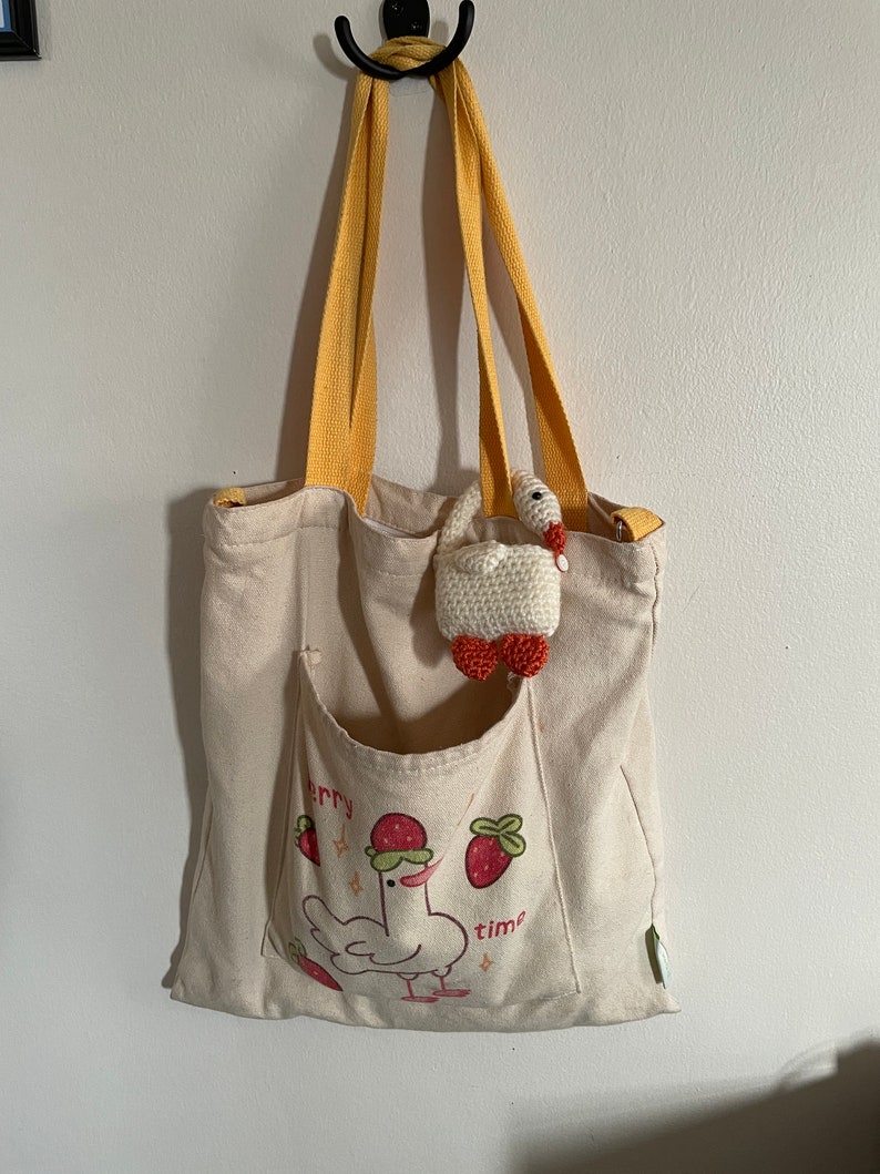 Pu&ograve; includere: Una borsa tote in tela beige con cinturini gialli. La borsa ha una tasca frontale con un'illustrazione di un pollo che indossa un cappello di fragola e il testo "berry time". Un piccolo anatroccolo bianco all'uncinetto con dettagli rossi &egrave; appeso alla borsa.