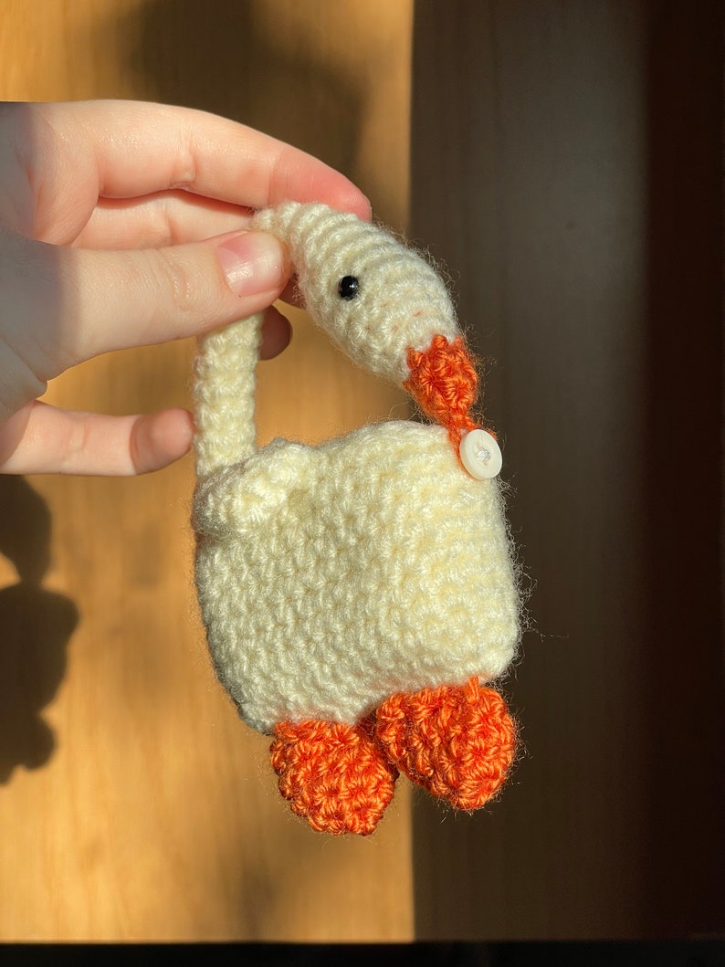 Pu&ograve; includere: Un'oca bianca all'uncinetto con piedi arancioni e un occhio a bottone nero. L'oca &egrave; tenuta da una mano.