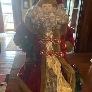 May include: A decorative Santa Claus figure, featuring a red velvet coat with faux fur trim and a matching hat. The figure has a white beard and holds a small Christmas tree. Gold and red accents adorn the coat.