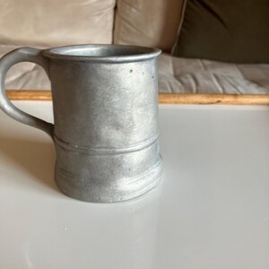 May include: A silver pewter beer stein with a handle. The stein is on a white surface.