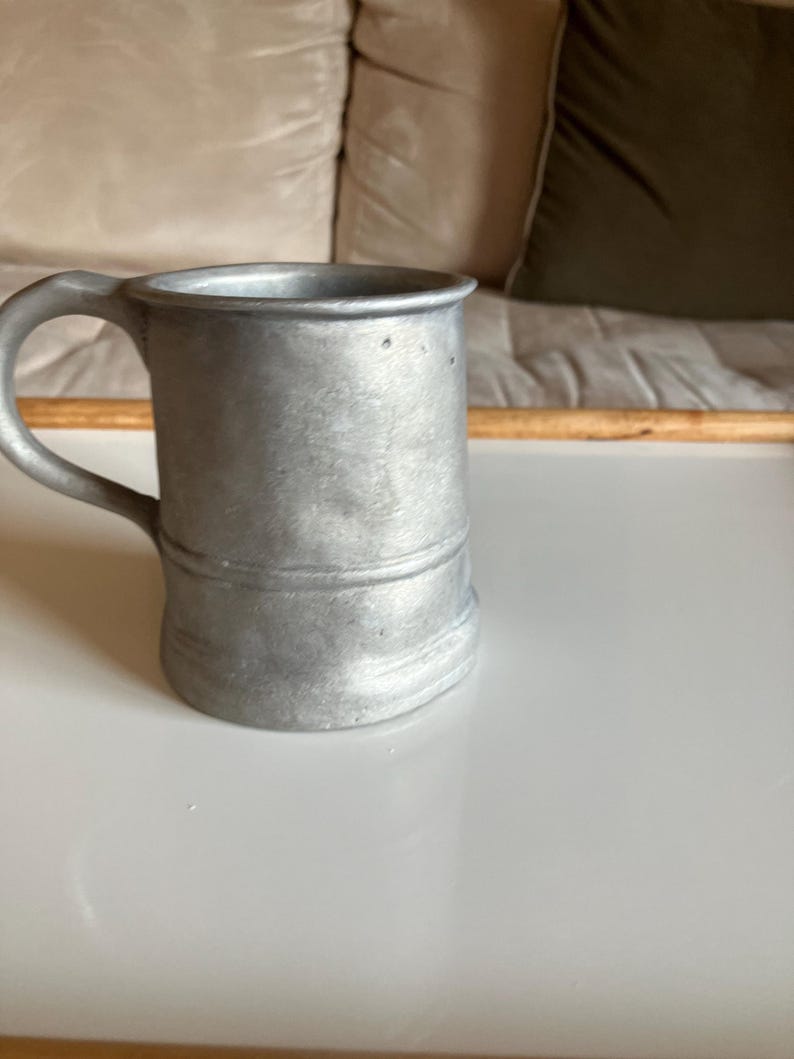 May include: A silver pewter beer stein with a handle. The stein is on a white surface.