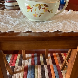 May include: A vintage white teapot with a gold trim and a floral pattern in shades of orange, yellow, and pink. The teapot is sitting on a white lace tablecloth on a wooden table.
