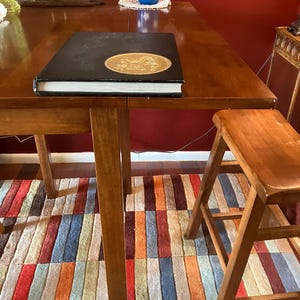 May include: A black hardcover book with a gold seal on the cover sits on a wooden table with a colorful striped rug underneath. A wooden stool is to the right of the table.