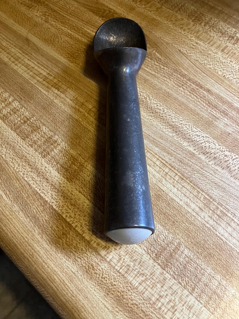 May include: A vintage metal ice cream scoop with a white plastic handle. The scoop has a worn, aged look and is perfect for serving ice cream or frozen desserts.
