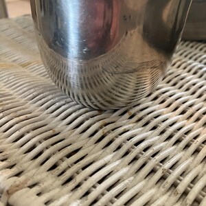 May include: A silver metal cup with a rounded bottom and a smooth, reflective surface. The cup is sitting on a white wicker surface.