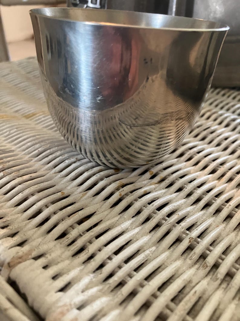 May include: A silver metal cup with a rounded bottom and a smooth, reflective surface. The cup is sitting on a white wicker surface.