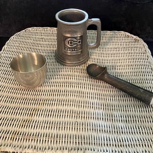 May include: A silver metal mug with a black and white logo that says "Control Cutler-Hammer" on a white wicker table with a silver metal cup and a silver metal ice cream scoop.