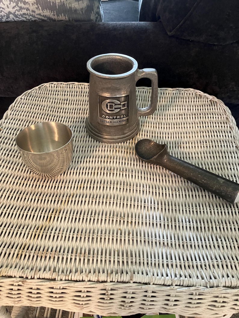 May include: A silver metal mug with a black and white logo that says "Control Cutler-Hammer" on a white wicker table with a silver metal cup and a silver metal ice cream scoop.