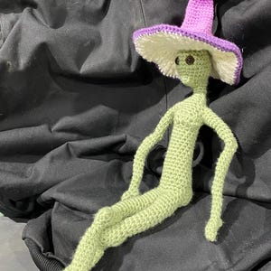 May include: A handmade crochet doll in the shape of an alien. The doll is light green with a purple and white hat. The doll has brown eyes and is sitting with its legs crossed. The doll is approximately 12 inches tall.