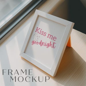 May include: White picture frame with a white mount and a pink inscription that reads "Kiss me goodnight" with a crescent moon and stars. The frame is sitting on a light wood surface.