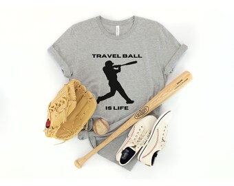 May include: A gray t-shirt with the text "Travel Ball is Life" and a silhouette of a baseball player swinging a bat. A baseball glove, a baseball, and a wooden baseball bat are also pictured.