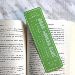 May include: A green bookmark shaped like a ticket with the words "Book Lovers Club" printed in white. The bookmark is on top of an open book. The text "Admit One" and "Cost: Free" is also printed on the bookmark.