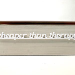 Peut inclure: Un marque-page blanc avec l'inscription "cheaper than therapy" en écriture cursive. Le marque-page est placé entre les pages d'un livre à couverture claire.