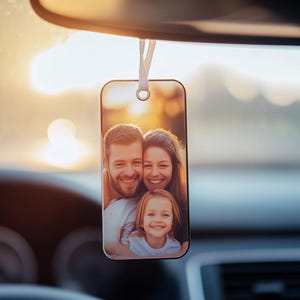 May include: A rectangular car air freshener hanging from a rearview mirror, featuring a printed photograph of a smiling family of three. The image shows a man, a woman, and a young child with a warm, golden sunset background and a white hanging string.