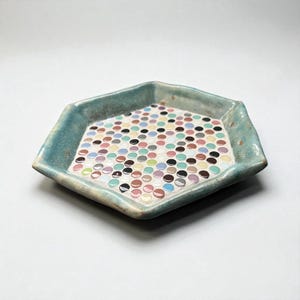 May include: A handmade ceramic tray with a hexagonal shape and a turquoise rim. The interior is decorated with a mosaic of small, colorful circles in various shades, including pink, blue, and black. This decorative tray is perfect for holding small items.