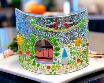 May include: A glass mosaic art piece depicting a whimsical garden scene with a small house, birds, flowers, and a gnome. The piece is made with colorful glass tiles and has a textured surface.