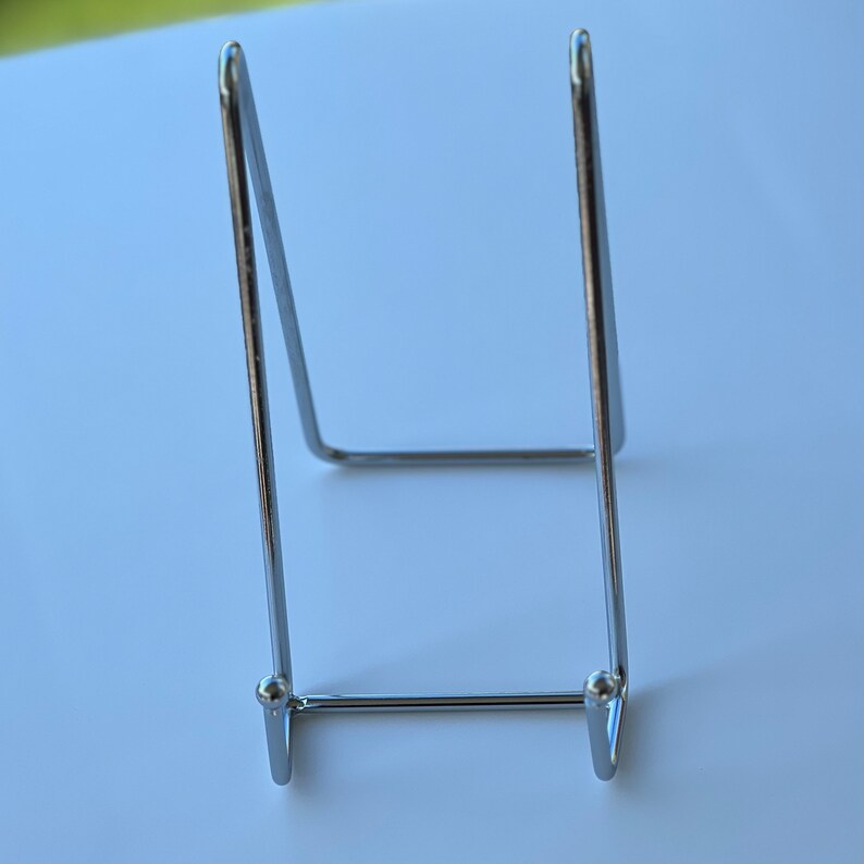 May include: A silver metal display stand for a picture or small object. The stand has a triangular shape with a flat base and two curved arms.