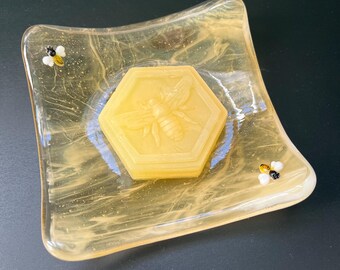 May include: A yellow glass soap dish with a honeycomb design and a bee-shaped soap bar. The dish has a textured surface and two small bee figurines.
