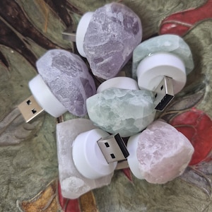 May include: A selection of USB flash drives, each with a distinctive design. They feature natural, irregularly shaped stones in purple, green, and pink, mounted on a white base. The drives are displayed in an ornate bowl.