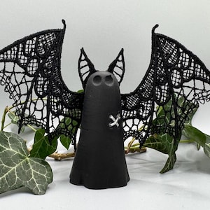 May include: A black bat ornament with delicate lace wings. The bat features a stitched 'X' detail and stands against a backdrop of green ivy. This Halloween decoration has a gothic aesthetic. The wings are made of lace, adding a touch of elegance to the spooky design.