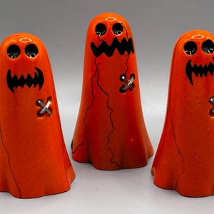 Peut inclure: Trois figurines fantômes orange avec des traits noirs et des accents argentés en forme de croix. Les fantômes ont une bouche en zigzag et deux yeux ronds. Décoration sur le thème d'Halloween.
