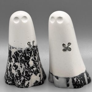 May include: Two white ghost-shaped ornaments with black and white patterned bases. Each has two eye indents and a metal cross detail. The ghosts stand side by side against a grey background.