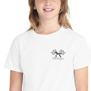 May include: White t-shirt featuring a small graphic of crossed checkered racing flags with a black ribbon and the name "Mackenzie" in script. The shirt is a classic crew neck style.