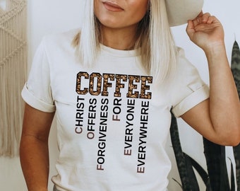 Op de afbeelding: Een wit T-shirt met het woord "COFFEE" in bruine en gouden glitterletters. De letters zijn omgeven door kleinere bruine en gouden glitterletters die "CHRIST OFFERS FORGIVENESS FOR EVERYONE EVERYWHERE" spellen.