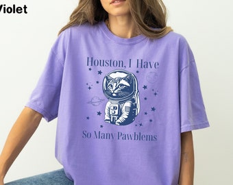 May include: A purple t-shirt with a graphic of a cat wearing an astronaut helmet and the text "Houston, I Have So Many Pawblems".