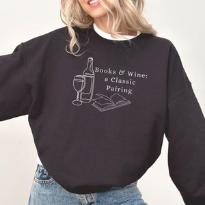 May include: Black sweatshirt with a white graphic design of a wine bottle, wine glass, and open book with the text "Books & Wine: a Classic Pairing".